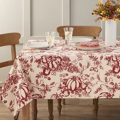 A table is adorned with a fabric featuring red botanical and pumpkin motifs, accompanied by glassware and a floral centerpiece.