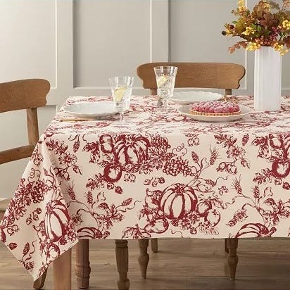 A table is adorned with a fabric featuring red botanical and pumpkin motifs, accompanied by glassware and a floral centerpiece.