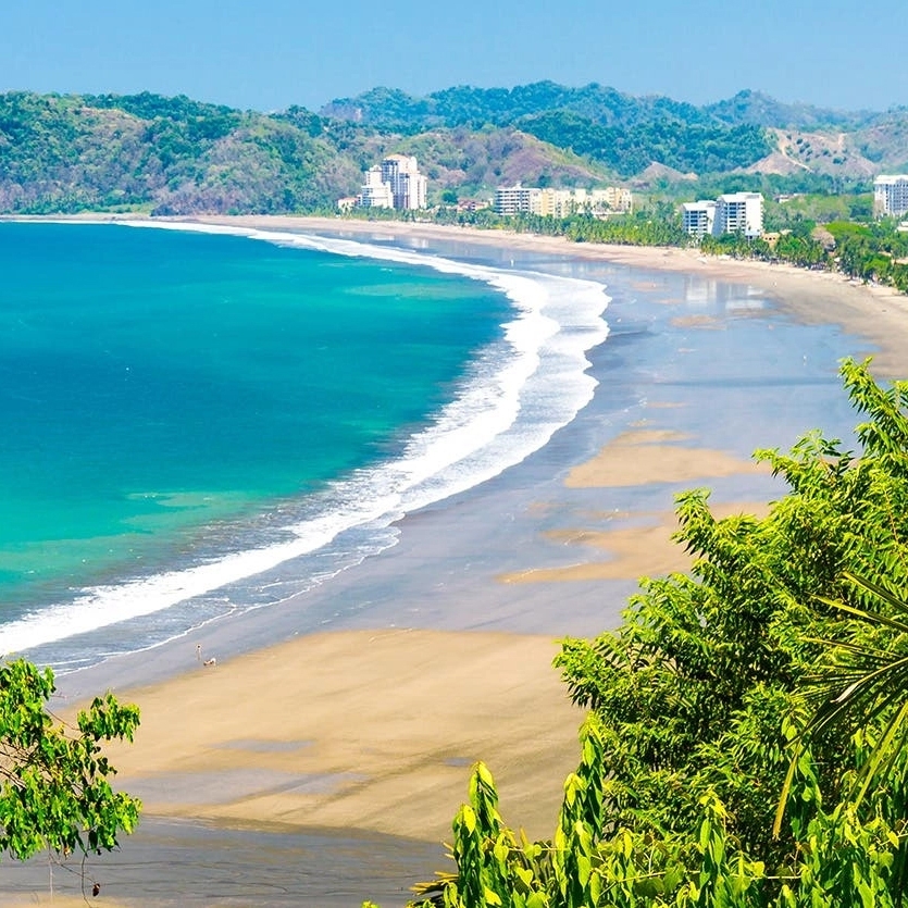 A stunning view of a crescent-shaped beach with clear blue-green water, bordered by lush greenery and a backdrop of buildings and hills.