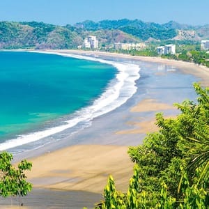 A stunning view of a crescent-shaped beach with clear blue-green water, bordered by lush greenery and a backdrop of buildings and hills.