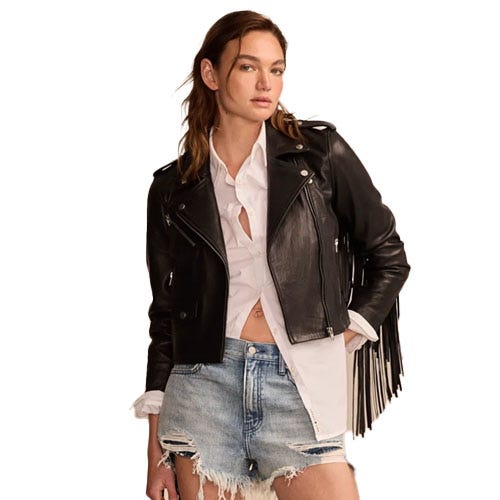 A woman is wearing a black leather jacket with fringe details, a white button-up shirt, and distressed denim shorts.