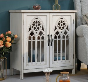 A white wooden sideboard with gothic-arch glass doors, accompanied by decorative items including a vase of roses and a gold-toned bottle.
