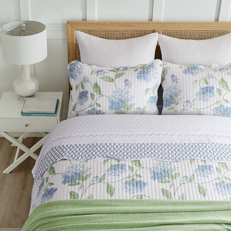 A quilted bed set with a blue and green hydrangea pattern and coordinating geometric designs on the borders flanked by white pillows against a wicker headboard.