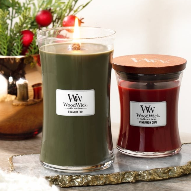 Two WoodWick candles are shown: one is tall and green labeled \“Frasier Fir,\“ with its flame lit, and the other is smaller, red, and labeled \“Cinnamon Chai,\“ with a wooden lid. They are set on a decorative surface with festive greenery in the background.