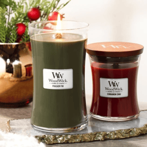 Two WoodWick candles are shown: one is tall and green labeled \“Frasier Fir,\“ with its flame lit, and the other is smaller, red, and labeled \“Cinnamon Chai,\“ with a wooden lid. They are set on a decorative surface with festive greenery in the background.