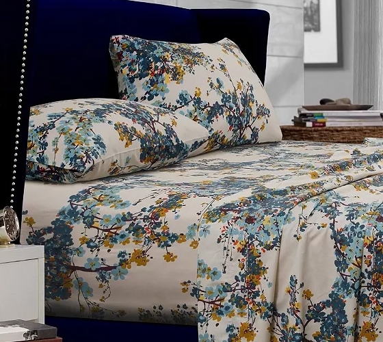 A bed with floral-patterned sheets and pillowcases in shades of blue, yellow, and green. The bedding is set against a dark blue upholstered headboard, and a nightstand with books is visible nearby.