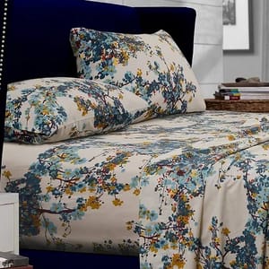 A bed with floral-patterned sheets and pillowcases in shades of blue, yellow, and green. The bedding is set against a dark blue upholstered headboard, and a nightstand with books is visible nearby.