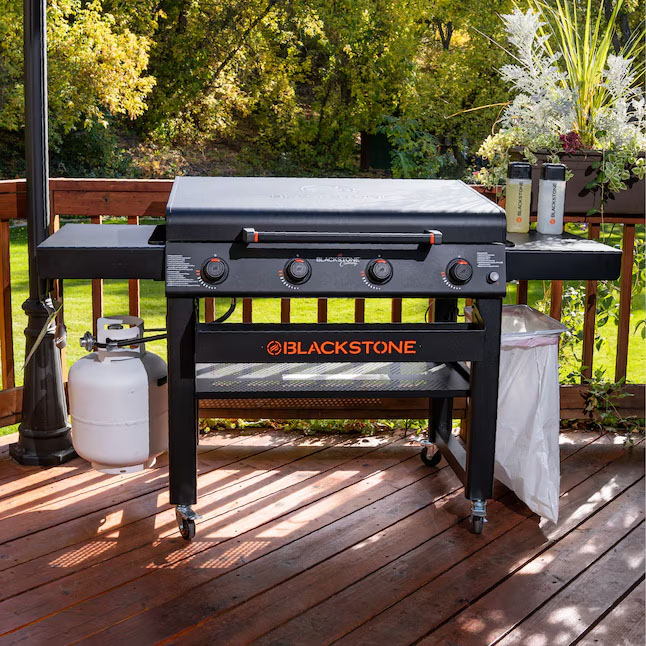 The 36\“ Blackstone Griddle with Hood is a propane flat-top grill featuring four control knobs, a storage shelf, a side shelf with two bottles, a trash bag, and is set on a wooden deck.