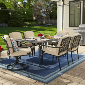 7-piece Hampton Bay Dining Set featuring a rectangular table and six cushioned chairs with beige upholstery. Includes swivel rockers, lattice chair backs, decorative pillows, and is set on a blue outdoor rug; patio string lights and a grill are visible.