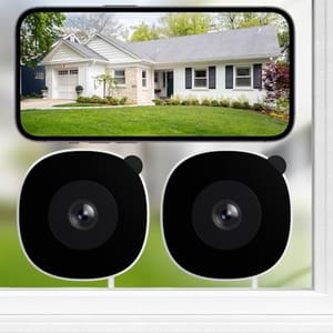 Two black surveillance cameras shown below a smartphone displaying a live feed of a house exterior with a lawn.