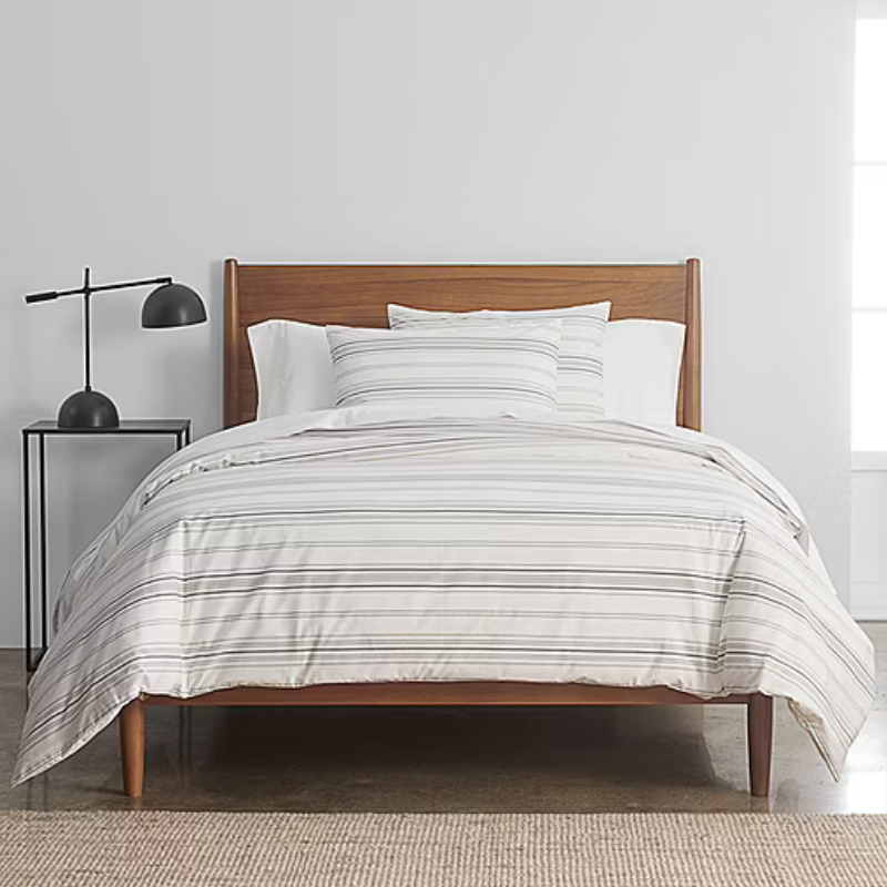 A bed with a wooden headboard features striped bedding set. A black table lamp sits on a small black side table next to the bed, all on a woven rug in a minimalist room.