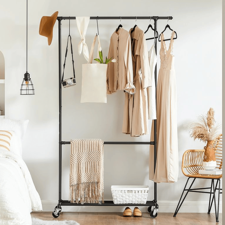 Clothing rack on wheels holding beige garments, a tote, and a camera. There's a hat, pendant lamp, woven chair with pampas grass, basket on the floor, slippers, and a folded blanket. Bedroom view includes a bed with white linens and a striped pillow.