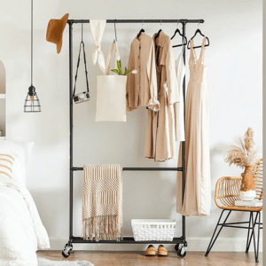 Clothing rack on wheels holding beige garments, a tote, and a camera. There's a hat, pendant lamp, woven chair with pampas grass, basket on the floor, slippers, and a folded blanket. Bedroom view includes a bed with white linens and a striped pillow.