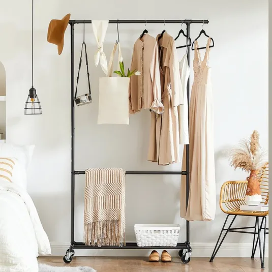 Clothing rack on wheels holding beige garments, a tote, and a camera. There's a hat, pendant lamp, woven chair with pampas grass, basket on the floor, slippers, and a folded blanket. Bedroom view includes a bed with white linens and a striped pillow.