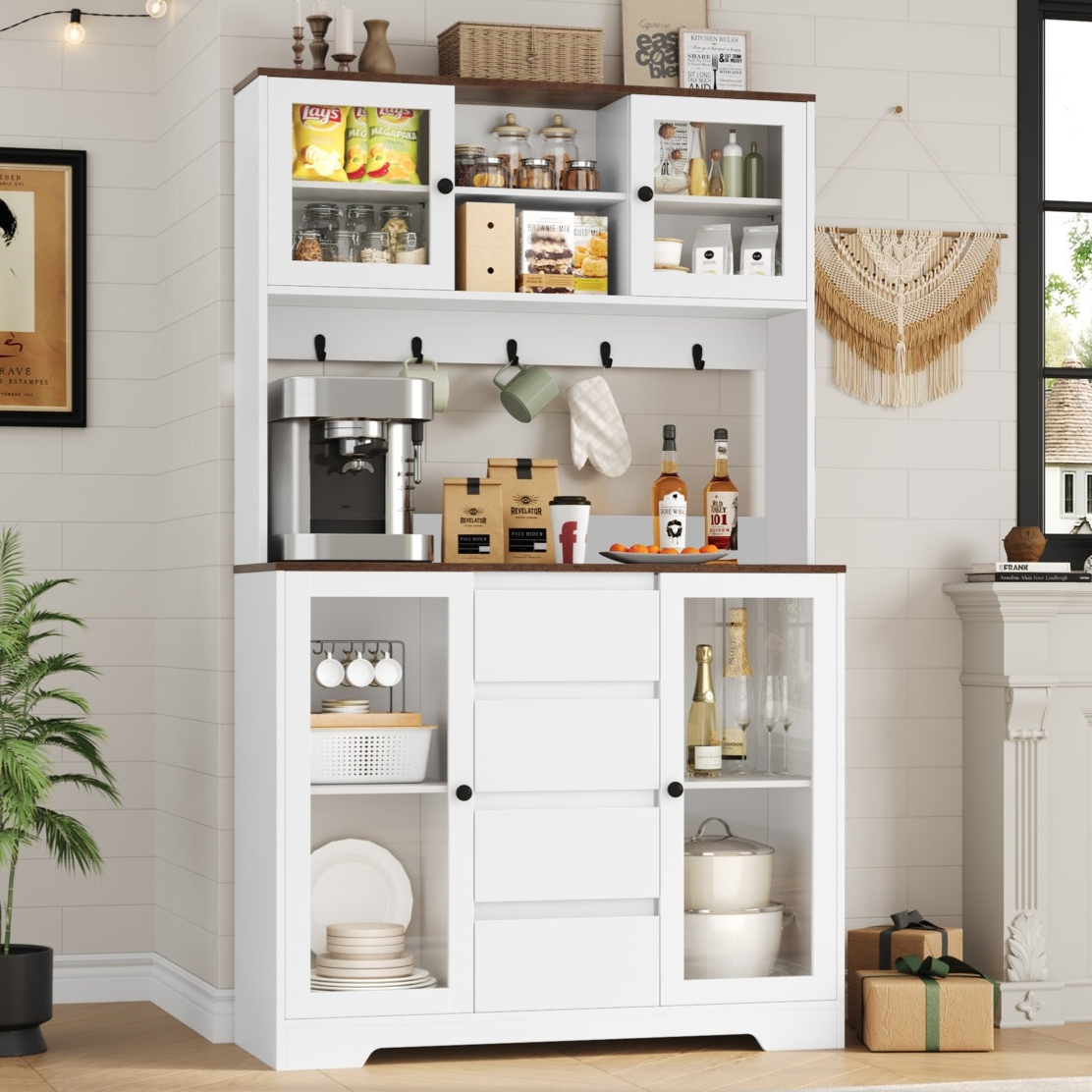 White kitchen hutch cabinet with dark wood accents, holding a coffee maker, various jars, snacks, condiments, mugs, and liquor bottles. Shelves display dishes, glasses, and pots. Boho wall hanging and plants add decor.