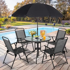 6-piece patio set includes a round glass-top table, four folding chairs, and a black umbrella. The table holds a vase of flowers, a jug of orange juice, books, and fruit, set on a wooden deck by a pool.