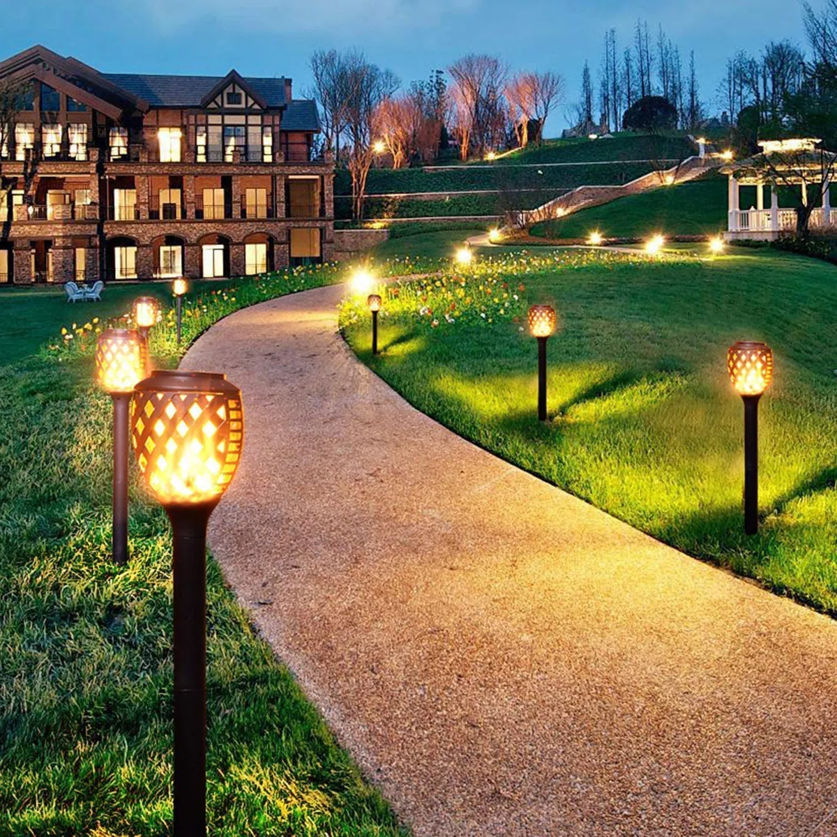 The photo showcases a pathway lined with decorative outdoor solar lamps, featuring a patterned design that emits warm light, enhancing the landscape of a large building's garden area during twilight.