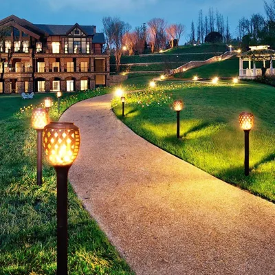 The photo showcases a pathway lined with decorative outdoor solar lamps, featuring a patterned design that emits warm light, enhancing the landscape of a large building's garden area during twilight.