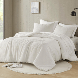 The King Boho Waffle Weave Comforter Set features a textured white comforter with matching pillow shams on a minimalist bed. It includes a round, fringed rug beneath the bed in a neutral, modern setting.
