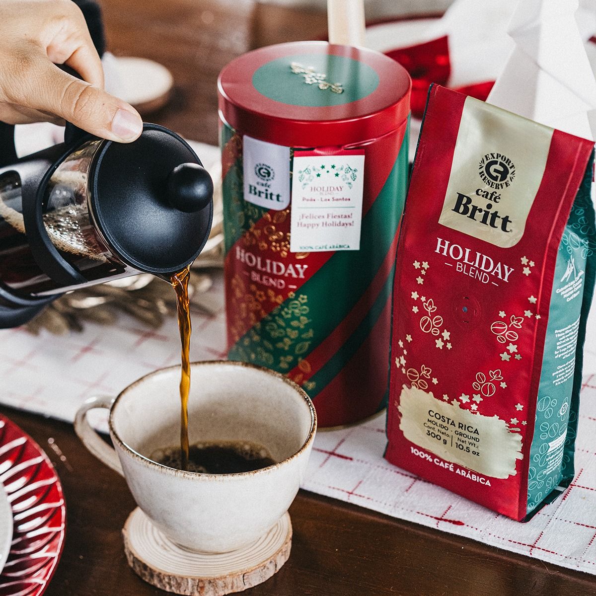A French press pours coffee into a cup beside Café Britt Holiday Blend coffee packaging, showcasing festive red and green designs.