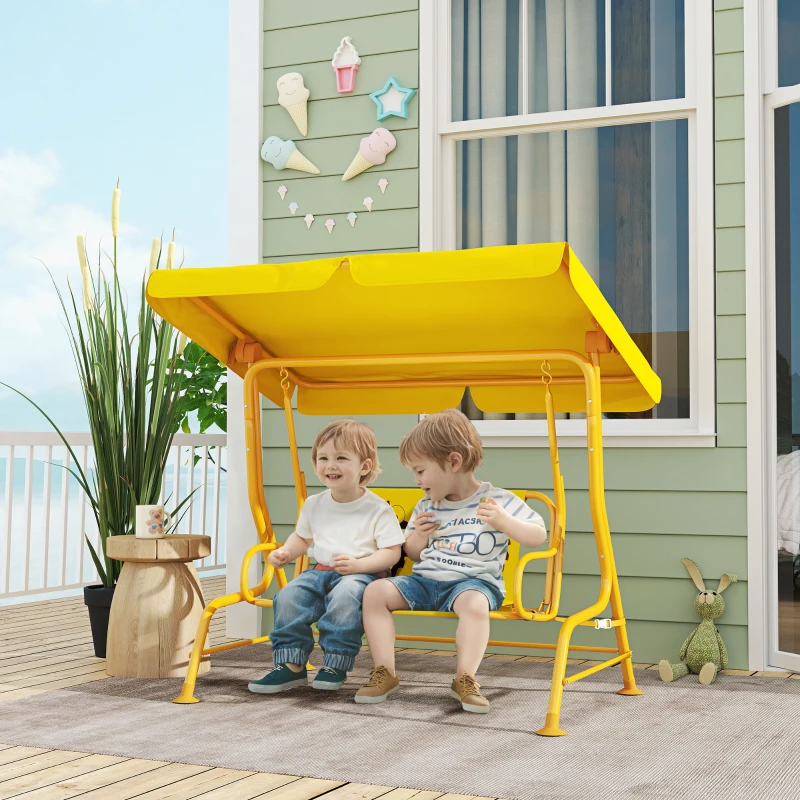 Two children sit on a yellow outdoor swing with a canopy. The setting includes a decorative wall with ice cream and star accents, a plant, a small wooden table, and a plush toy rabbit.