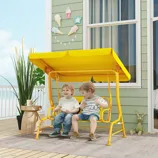 Two children sit on a yellow outdoor swing with a canopy. The setting includes a decorative wall with ice cream and star accents, a plant, a small wooden table, and a plush toy rabbit.