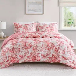 A floral-patterned comforter set with red and white colors is displayed on a bed, accompanied by matching pillow shams. The room features a wood nightstand with a lamp, wall art, and a window with a view.