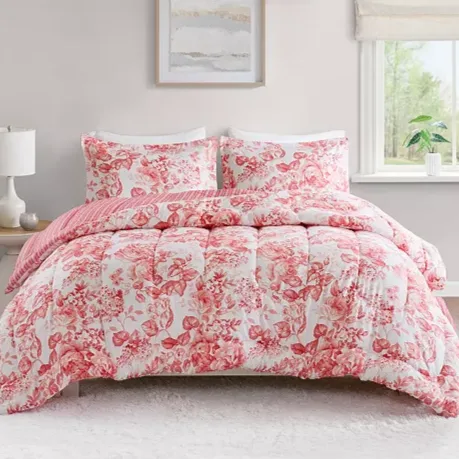 A floral-patterned comforter set with red and white colors is displayed on a bed, accompanied by matching pillow shams. The room features a wood nightstand with a lamp, wall art, and a window with a view.