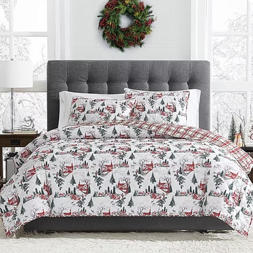 A cozy bedroom scene with winter-themed bedding featuring red cabins and trees, a gray tufted headboard, neutral side tables, and a festive wreath on the white-paneled wall.