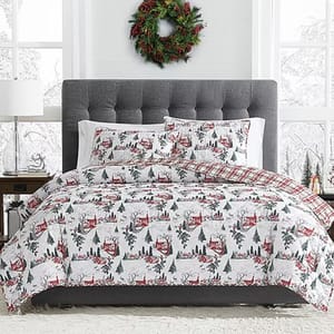 A cozy bedroom scene with winter-themed bedding featuring red cabins and trees, a gray tufted headboard, neutral side tables, and a festive wreath on the white-paneled wall.