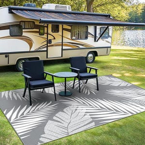 A recreational vehicle with an extended awning is parked by a lake, accompanied by two black chairs and a small round table set on a rug with a white leaf pattern.