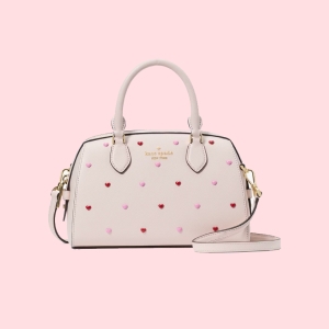 A cream-colored handbag adorned with small red and pink heart motifs, featuring a top handle and a detachable strap.