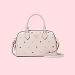A cream-colored handbag adorned with small red and pink heart motifs, featuring a top handle and a detachable strap.