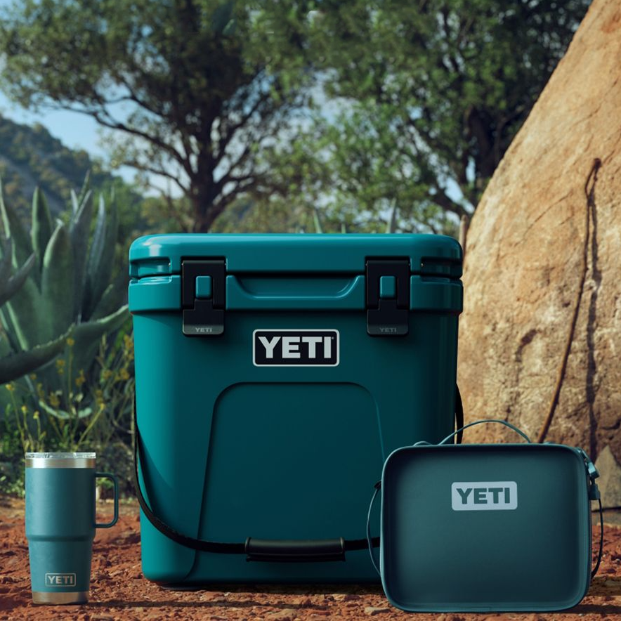 A YETI cooler, tumbler, and lunch box in teal color, set outdoors near a cactus and a tree.