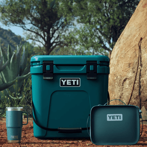 A YETI cooler, tumbler, and lunch box in teal color, set outdoors near a cactus and a tree.