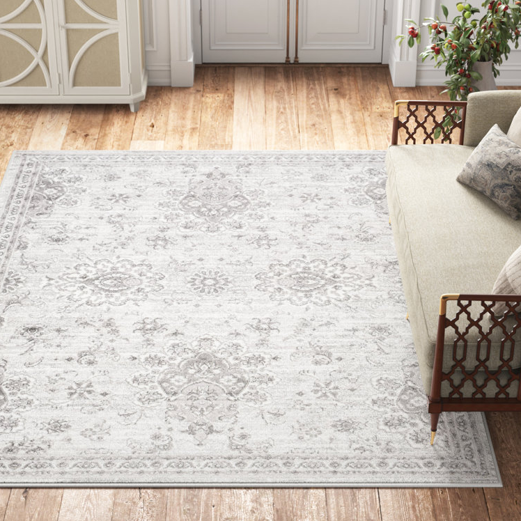 The Kelly Clarkson Ingrid Rug features a 5' x 7' size with a delicate, vintage grey floral pattern on a light background, resting on a wooden floor next to a light-colored sofa and a decorative cabinet.