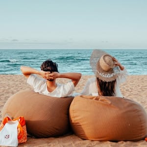 Two people relax on brown bean bags facing the ocean with a beach bag nearby. One wears a sun hat, suggesting a casual and comfortable beach setting.