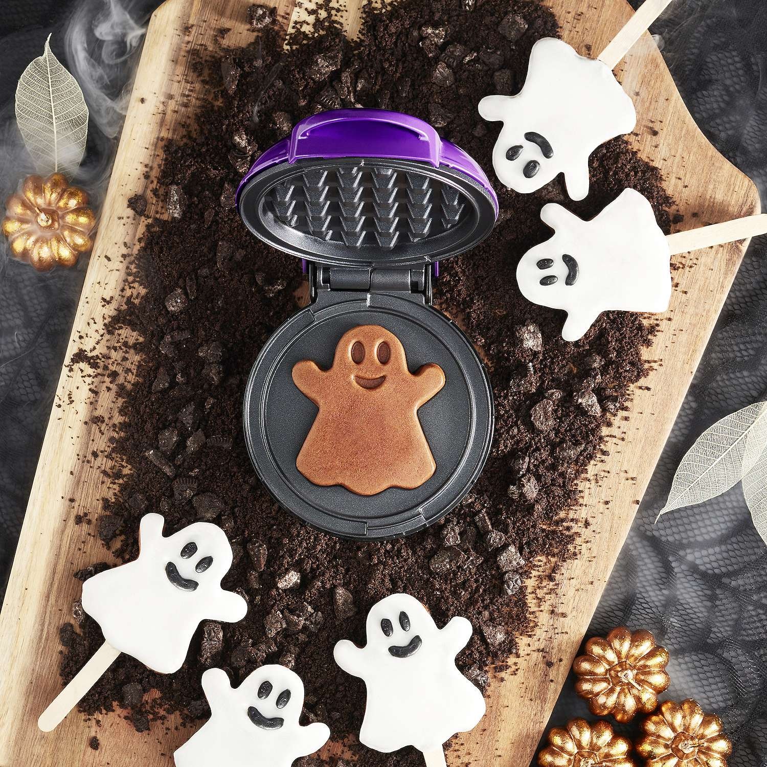 Waffle maker with a ghost-shaped mold, accompanied by ghost-shaped popsicles and decorative pumpkins on a wooden surface.