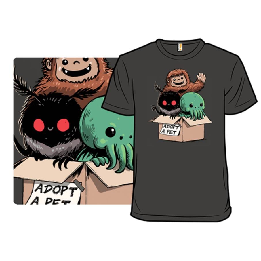 A black T-shirt features cute cartoon monsters, including a furry creature, an octopus-like figure, and a shadowy being with red eyes, all sitting in a cardboard box labeled \“Adopt A Pet.\“