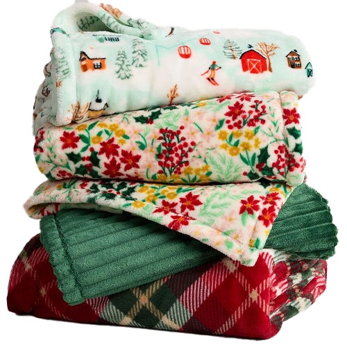 A stack of cozy, patterned blankets featuring holiday-themed designs, including winter scenes, floral prints, and plaid patterns, in various colors and textures.