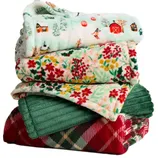 A stack of cozy, patterned blankets featuring holiday-themed designs, including winter scenes, floral prints, and plaid patterns, in various colors and textures.