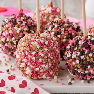 Set of six caramel apples coated with pink, white, and chocolate sprinkles, heart-shaped candies, and nuts, each on a wooden stick, displayed on a white surface with a heart-patterned cloth.