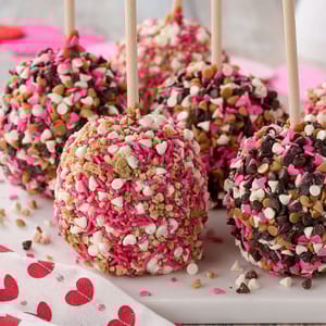 Set of six caramel apples coated with pink, white, and chocolate sprinkles, heart-shaped candies, and nuts, each on a wooden stick, displayed on a white surface with a heart-patterned cloth.