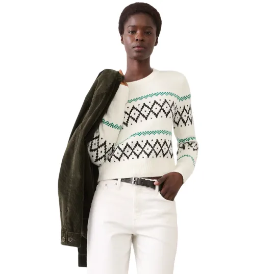 A person wears a white sweater with a black and green geometric pattern, paired with white pants and a black belt, holding an olive green jacket over one shoulder, and standing next to a black chair.