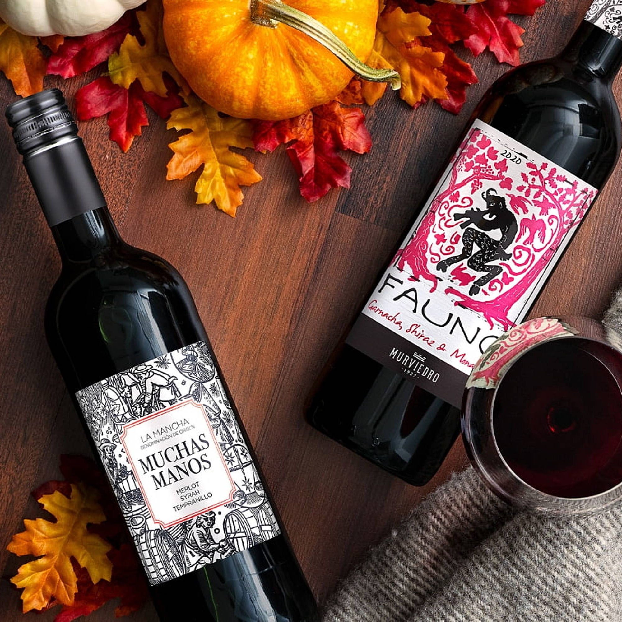 Two bottles of red wine are displayed among autumn leaves and a pumpkin, with one bottle opened and a glass of red wine alongside.