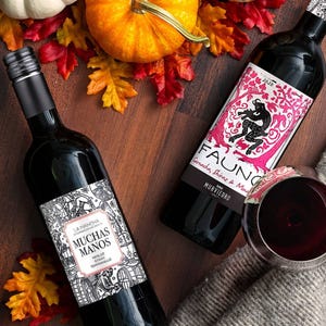 Two bottles of red wine are displayed among autumn leaves and a pumpkin, with one bottle opened and a glass of red wine alongside.