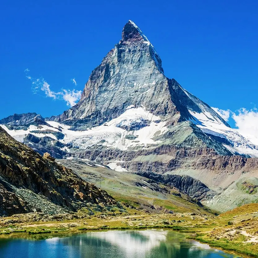 A majestic mountain peak, likely the Matterhorn, rises above a small tranquil lake, with snow patches on its slopes under a clear blue sky.