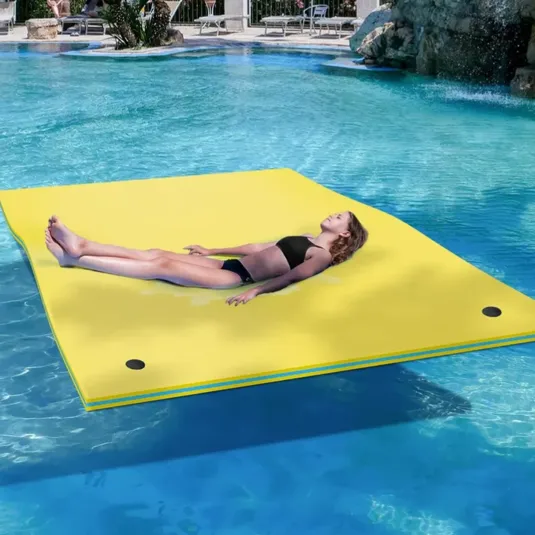 A person relaxes on a large yellow water mat floating in a pool, with surrounding lounge chairs and plants in the background.