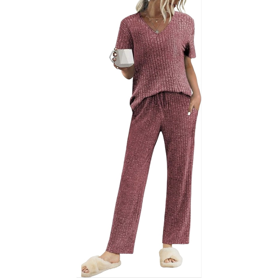 A woman wears a comfy maroon ribbed loungewear set featuring a short-sleeve V-neck top and matching drawstring pants, paired with fluffy cream slippers. She is holding a white mug.