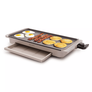 An electric griddle with a non-stick surface is cooking eggs, bacon, and pancakes, with an adjustable temperature dial and a slide-out drip tray.